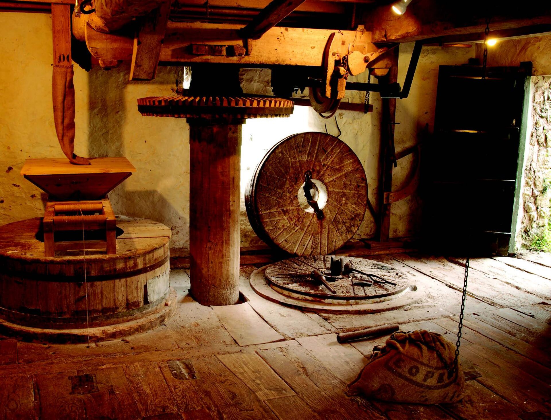 A darkly lit room insdie the mill with large stone mechanical wheel cogs going through a wooden floor.